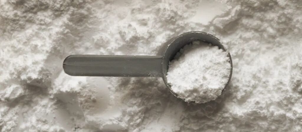 A large pile of a white powdered supplement with a grey plastic scoop centered in the frame.